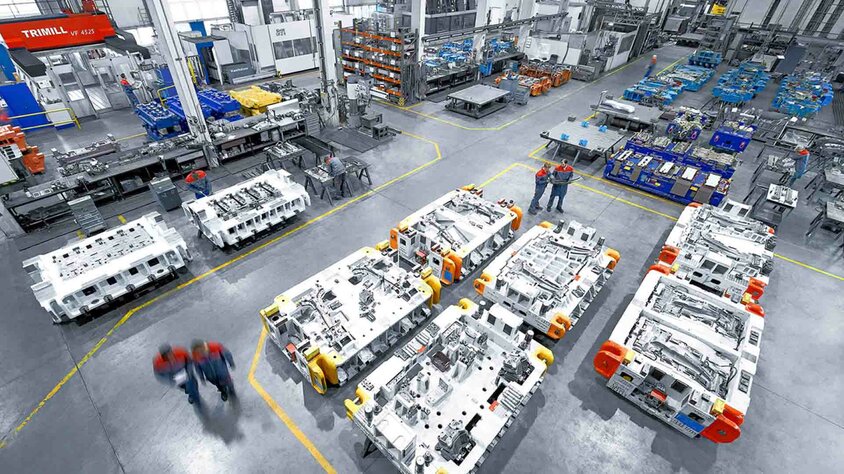 A Modern Tool Manufacturing Facility with Large stamping and pressing tools. The production hall is clean and well-organized, with several large tools positioned on the floor. In the background, various machines and workbenches can be seen, while in the foreground, two employees in work attire are engaged in conversation. This image highlights the professional and efficient use of tools and machinery in the tool manufacturing industry, specifically for the automotive sector.