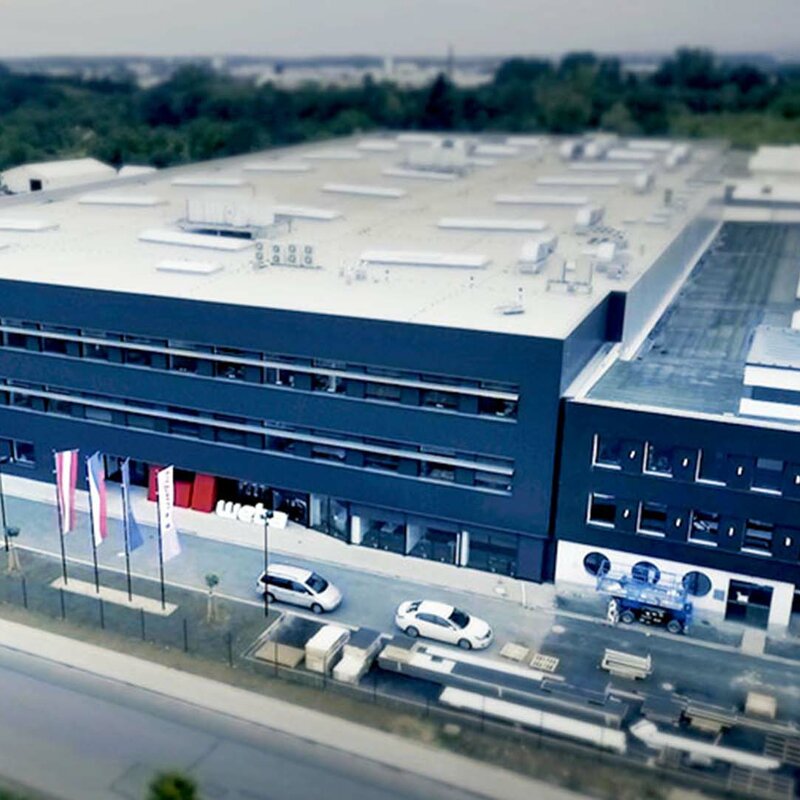 Aerial view of weba's production plant for hot forming and series production in Olomouc. The photo shows the anthracite-colored front of the building and a huge production hall behind it. The Austrian, Czech, European and company flags fly in front of the plant.