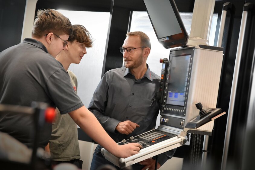 An apprenticeship trainer explains the control system of a machining center to two apprentices in the field of metal engineering, Module H8 Machining Technology. The apprenticeship at weba in Steyr combines practical experience with theoretical knowledge in machining technology.