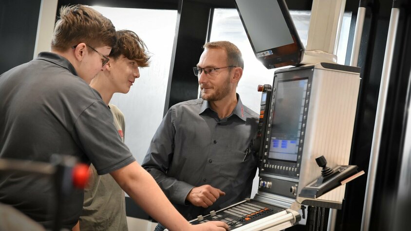 Ein Lehrlingsausbilder erklärt zwei Lehrlingen im Bereich Metalltechnik, Modul H8 Zerspanungstechnik, die Steuerung eines Bearbeitungszentrums. Die Lehre in Steyr bei weba kombiniert praktische Erfahrung mit theoretischen Know-how in der Zerspanungstechnik.