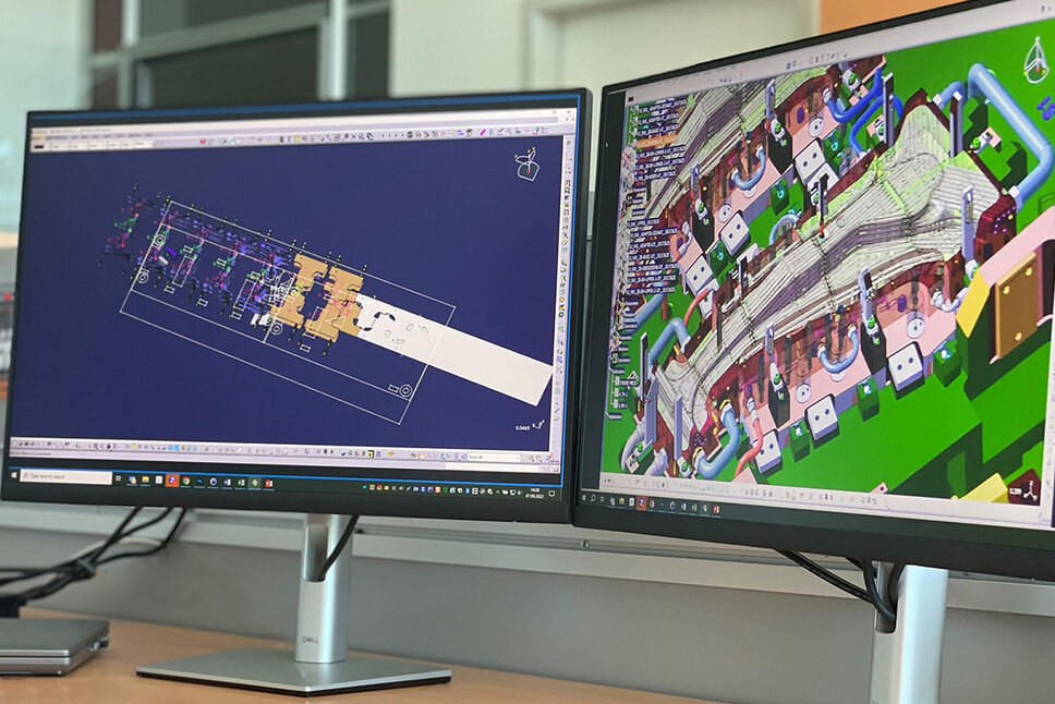 Two computer screens are displayed: one shows a detailed close-up of a CAD design for a hot forming tool used in the production of B-pillars, while the other screen depicts the transfer setup of a tool.