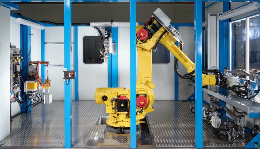 A modern joining technology facility features a yellow FANUC robotic arm positioned centrally in a shielded workspace, ready for automated adhesive tasks. Surrounded by blue frame structures, the scene showcases a high-tech manufacturing environment with precise mechanical components and advanced control systems. This facility represents precision, efficiency, and automation in joining technology.