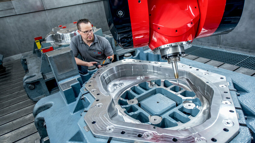 Ein Facharbeiter bedient eine hochmoderne CNC-Fräsmaschine mit einem großen Werkzeugbauteil. Die glänzende Metalloberfläche des Bauteils zeigt präzise Zerspanungsergebnisse, während die rote Fräseinheit prominent im Vordergrund zu sehen ist.