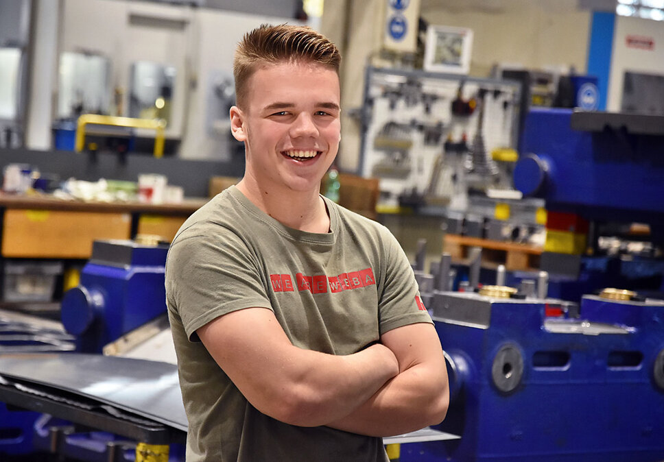 An apprentice at weba Werkzeugbau, photographed in the production hall, smiles directly at the camera. He is wearing a T-shirt that says 'We are WEBA,' expressing his pride and sense of belonging to the company. In the background, several workbenches and parts of a dark blue forming tool are visible, indicating the practical and intensive learning environment in which the apprentice is being trained. The image radiates youthfulness, optimism, and the apprentice's commitment to his training while highlighting the supportive and educational atmosphere at weba. The image symbolizes apprenticeship jobs in Steyr at weba Werkzeugbau.