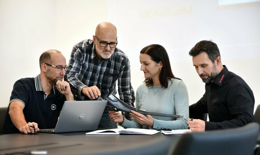  Three male employees and one female employee of weba Werkzeugbau are sitting in a meeting discussing a steel component. A laptop and documents are on the table.