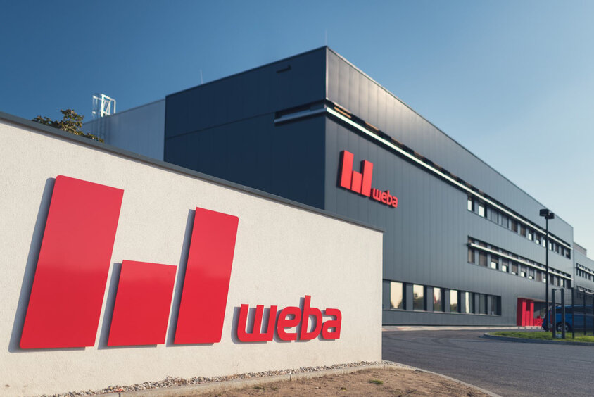 Das Bild zeigt die Außenansicht des Produktionsstandorts weba Olomouc. Die Gebäudefront ist anthrazitgrau gestaltet, was einen modernen und professionellen Eindruck vermittelt. Vor der Einfahrt ist das rote weba-Logo auf der Mauer deutlich sichtbar. Die Szene strahlt Stabilität und Markenidentität aus, während das Logo Unternehmen und Standort identifiziert.