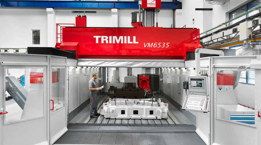 The picture shows a large Trimill machining center with the door open, allowing a view of the milling table. A large casting can be seen on the milling table, waiting to be machined. The scene conveys the impression of industrial production and precision 