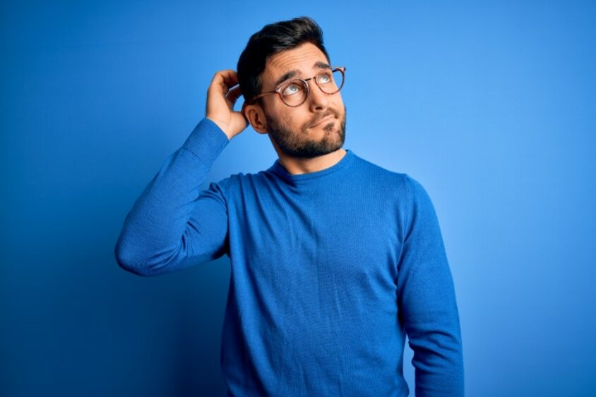 A man stands in front of a blue background, thoughtfully scratching his head and looking up, symbolizing frequently asked questions (FAQs) in the application process.