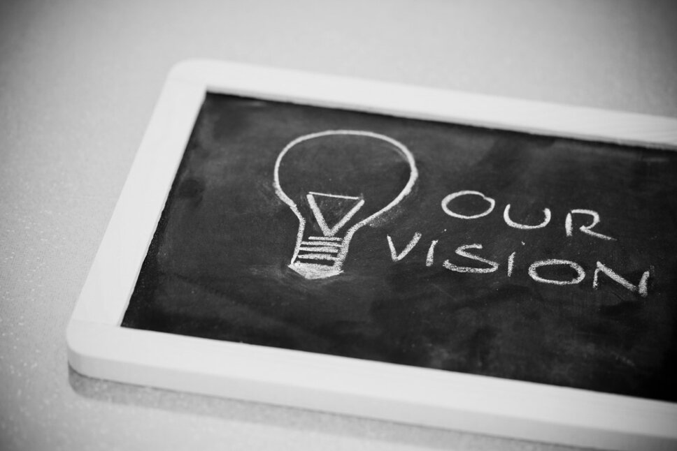 A chalkboard with a white frame lying on the table, featuring a chalk drawing of a light bulb and the text "Our Vision." The photo symbolizes an innovative work culture and outstanding career opportunities in Steyr.