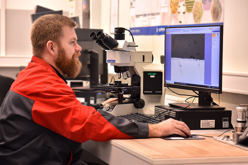 A quality assurance employee sits at a desk, intently looking at a computer screen displaying material testing results. In the background of the room, a microscope is visible, highlighting the importance of precise examinations in this field.