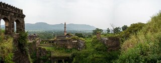 Eine historische Ruinenlandschaft mit üppiger Vegetation, einer alten Steinmauer im Vordergrund und einer hohen, markanten Säule im Hintergrund, eingebettet in eine weite grüne Landschaft.