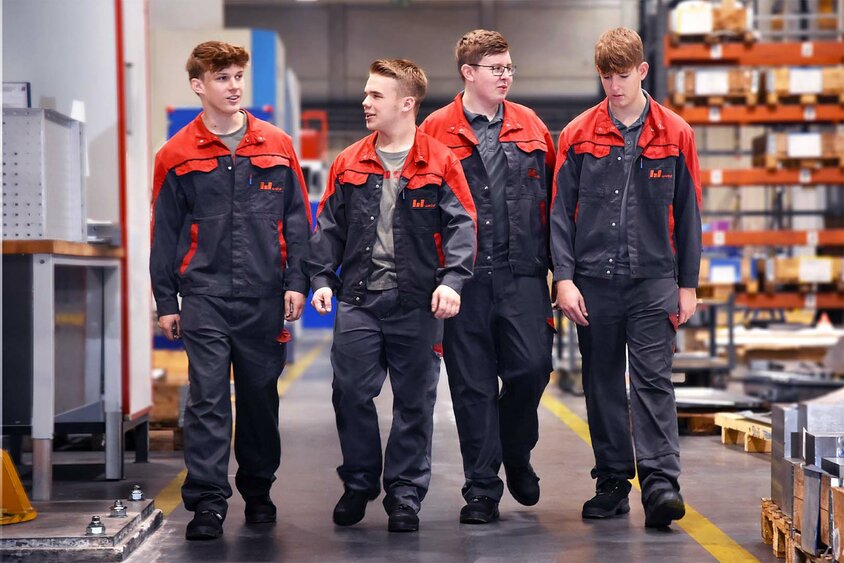 Four dedicated apprentices from Weba Werkzeugbau stroll through the lively production hall. They chat animatedly as they walk past impressive metal-cutting machines and well-stocked shelves of tools and materials. The picture symbolizes the apprenticeship in metal technology at weba and conveys an atmosphere of learning and practical application, embedded in the real working environment of a modern toolmaking company.