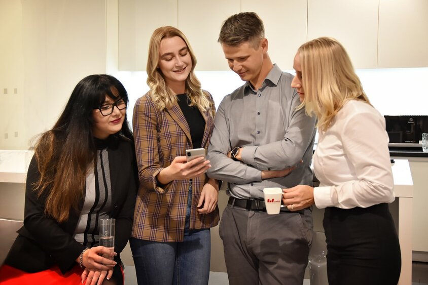 Three female employees and one male employee from weba Olomouc stand smiling in an office kitchen, collectively looking at a smartphone. Two of them are holding coffee cups, indicating a relaxed and collegial atmosphere.