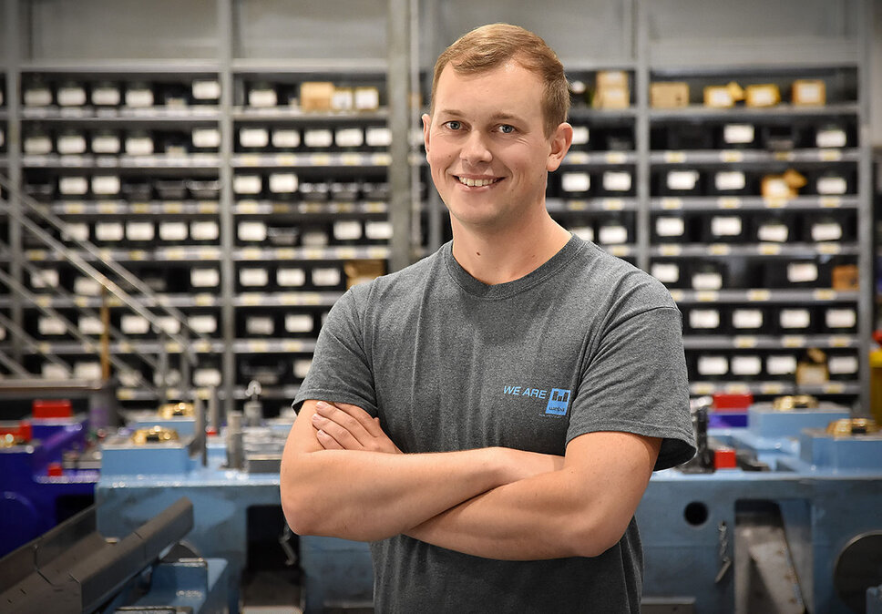 A tool engineer from weba Werkzeugbau, photographed in the production environment, looks directly and proudly at the camera with a smile. He stands in a confident pose that highlights his pride in his work. In the background, neatly arranged equipment shelves and a distinctive blue forming tool are visible, indicating a well-organized and technologically advanced work environment. The image conveys a sense of professionalism and satisfaction in the workplace, emphasizing the technician's personal connection to his work and workspace. The image symbolizes internship jobs in Steyr.