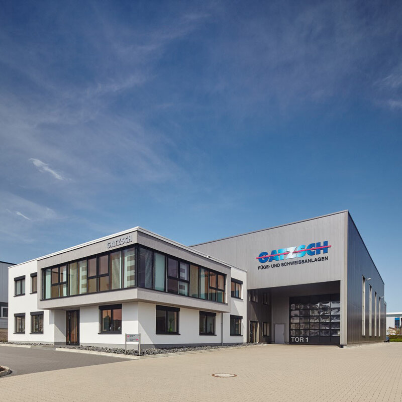 The exterior view of the Gatzsch Schweißtechnik GmbH plant shows a bright blue sky and the administration building in focus. Behind it is the production plant, on which a large logo with the words 'Gatzsch Füge- und Schweissanlagen' is clearly visible.