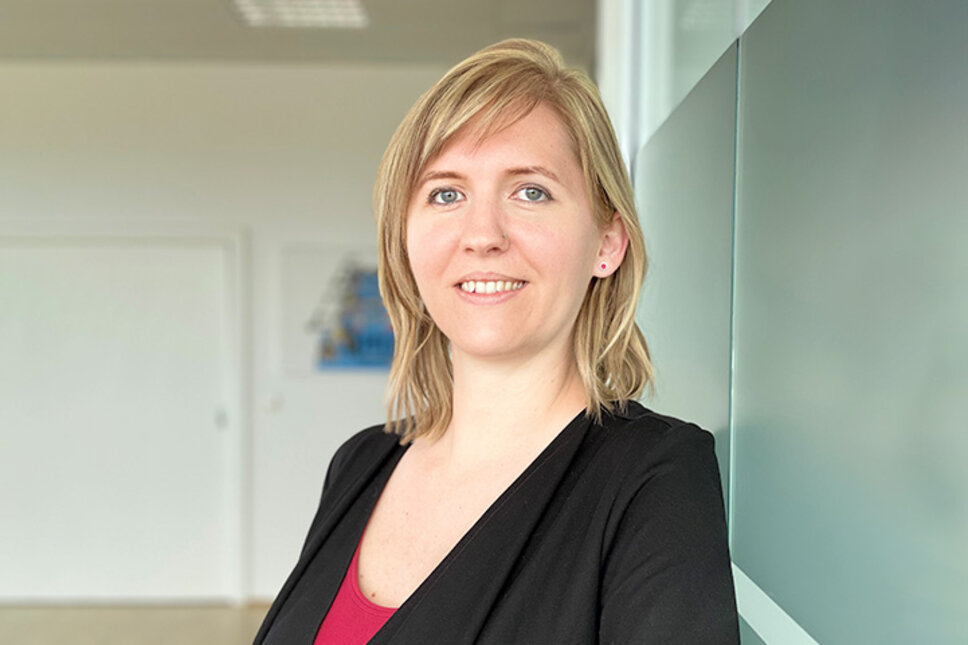 Portrait of Tanja Burghuber, HR Manager at weba Werkzeugbau, leaning relaxed against a frosted glass wall in Dietach, with a professional and friendly expression, emphasized by the soft lighting and blurred background. The photo is used in the contact section of weba Werkzeugbau's job vacancies in Steyr.