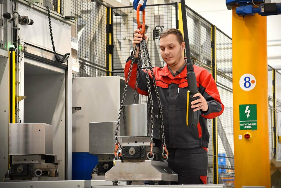 A machine operator is working at a setup station of an automated machining center for contract manufacturing. He is carefully placing tools and components into the setup positions. The focus is on the precise placement of precision tools in the designated positions on the complex machine bed. In the background, the industrial environment with advanced technology is visible.