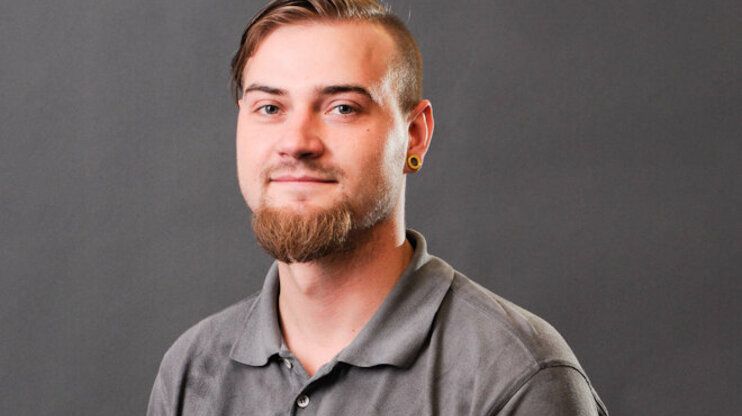 A portrait photo of Bronislav Škoda, a young man with short, side-shaved hair and a well-groomed full beard. He is wearing a gray polo shirt with the weba logo and is looking warmly into the camera. His ears are adorned with yellow plugs, and the background is a neutral gray studio backdrop.