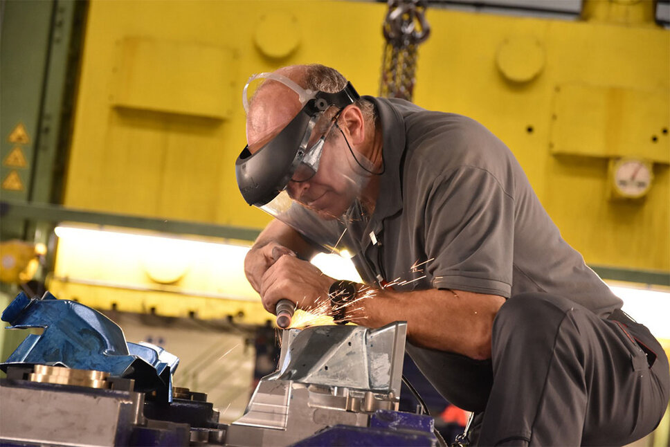 A toolmaking technician works on a tool insert in the forming press. During the grinding process, sparks fly around. The technician is equipped with a face shield that ensures his safety during the precise and demanding work process in the tool tryout.