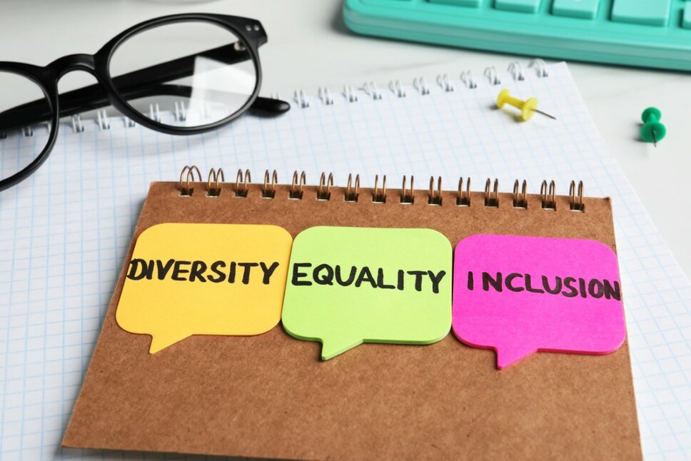 Backside of a notepad with three colorful post-its displaying the words "Diversity, Equality, and Inclusion." Next to it are a pair of black-rimmed glasses, a turquoise keyboard, and two push pins in green and yellow.