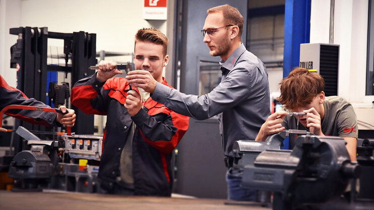 Ein Lehrling in der Werkzeugbautechnik, konzentriert beim Messen eines Stahlklotzes mit einem Messschieber, während der Ausbilder neben ihm steht und etwas erklärt und zeigt. Daneben spannt ein weiterer Lehrling aufmerksam einen Stahlklotz in einen Spannbock, was die praktische und lehrreiche Umgebung der Ausbildung verdeutlicht. Das Bild ist Symbolfoto für die Lehre in Steyr in der Werkzeugbautechnik Modul H6 aus der Metalltechnik bei weba Werkzeugbau.