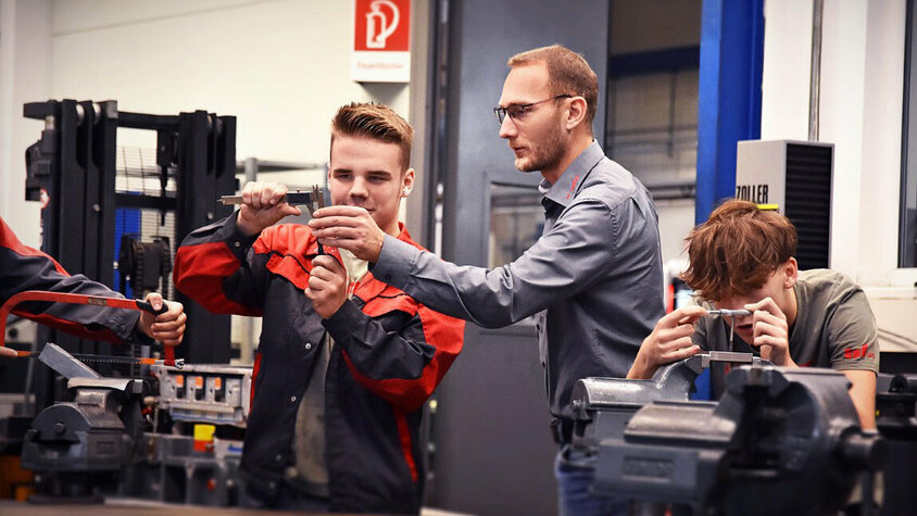 Ein Lehrling in der Werkzeugbautechnik, konzentriert beim Messen eines Stahlklotzes mit einem Messschieber, während der Ausbilder neben ihm steht und etwas erklärt und zeigt. Daneben spannt ein weiterer Lehrling aufmerksam einen Stahlklotz in einen Spannbock, was die praktische und lehrreiche Umgebung der Ausbildung verdeutlicht. Das Bild ist Symbolfoto für die Lehre in Steyr in der Werkzeugbautechnik Modul H6 aus der Metalltechnik bei weba Werkzeugbau.