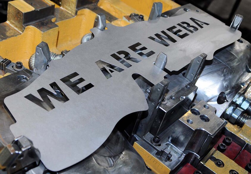  A blank of a stamped part is positioned in a forming tool. The inscription 'WE ARE WEBA' has been carefully laser-engraved into the blank, highlighting precise craftsmanship and the company's identity. This image symbolizes teamwork and passion and is used on the "Jobs in Steyr" page for experienced professionals and skilled workers.