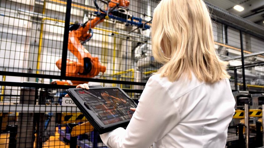 Eine Frau steht in einer industriellen Umgebung bei weba Werkzeugbau vor einem großen Robotergreifer einer Warmumformlinie. In ihren Händen hält sie ein Tablet, auf dem sie Daten überwacht oder Steuerbefehle für den Roboter eingibt. Der Fokus liegt auf der Interaktion zwischen moderner Technologie und menschlichem Eingreifen.