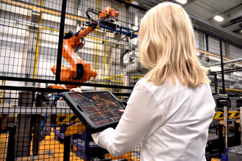 A woman stands in an industrial environment at weba Werkzeugbau in front of a large robotic gripper of a hot forming line. She holds a tablet in her hands, monitoring data or inputting commands for the robot. The focus is on the interaction between modern technology in tool manufacturing and parts production and human intervention.