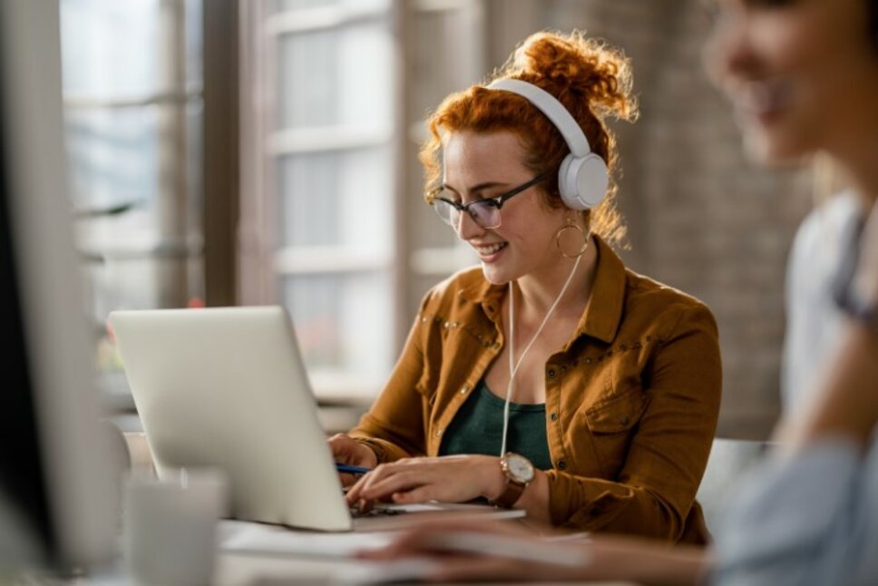 A smiling, creative young woman listens to music with headphones while working at a computer in the office during her internship job at weba in Steyr.