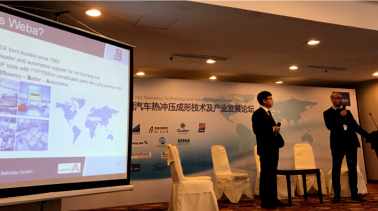 Auf der Bühne präsentieren zwei Vertriebsmitarbeiter von weba auf der Automotive Forum Messe in China  das Unternehmen und die Vorteile der Warmumformung für den Leichtbau in der Automobilindustrie.