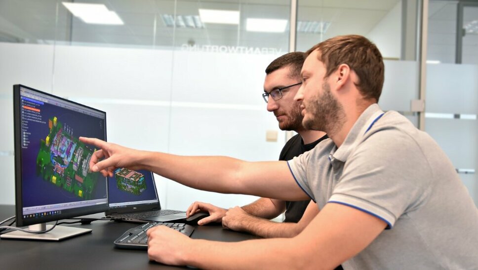 Two people are sitting in front of two computer screens on which a CATIA file with a tool design is open. One of the people points to a specific detail of the tool on the screen while they are apparently discussing the design.