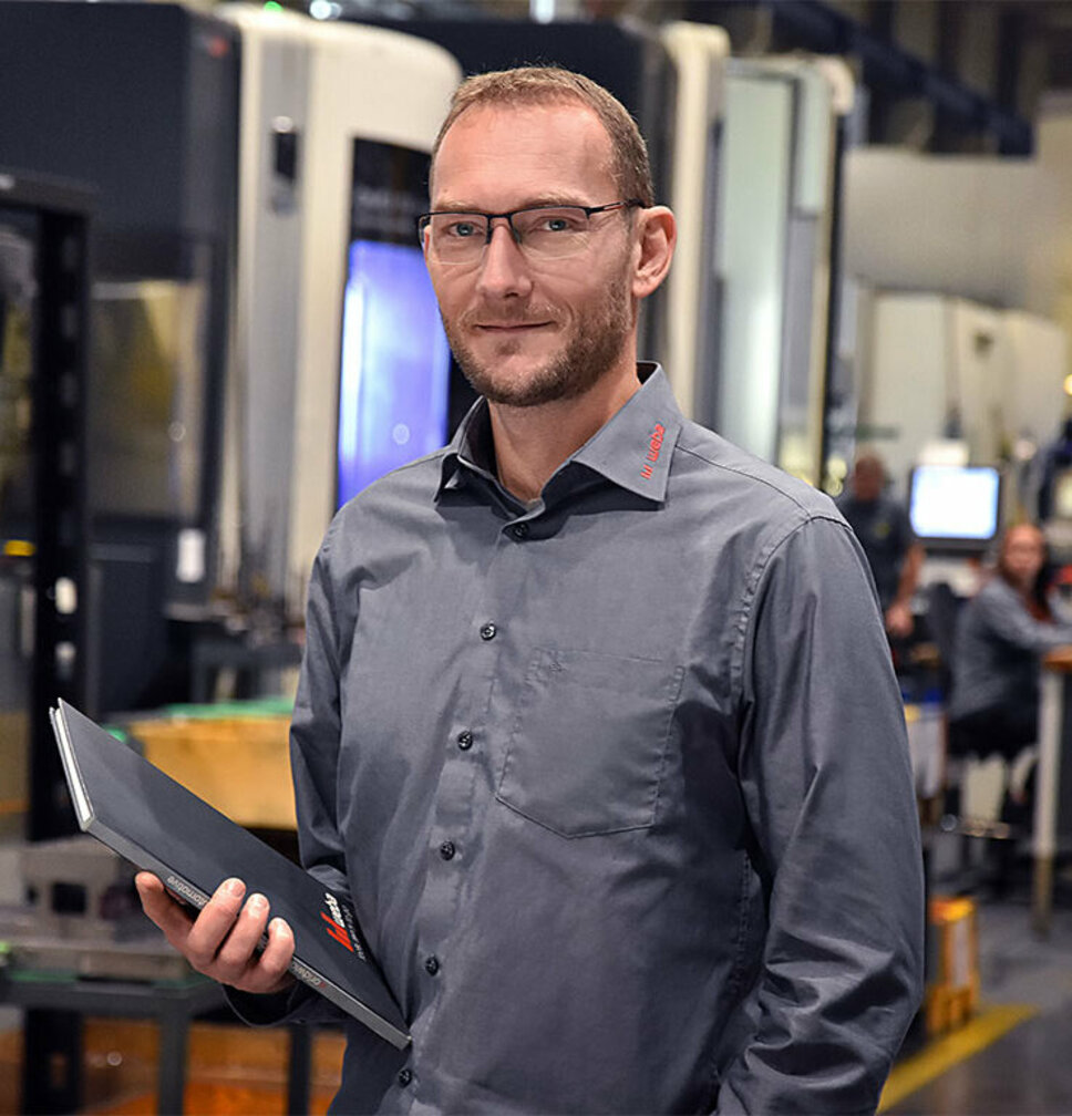 Portrait photo of the apprenticeship trainer at weba Werkzeugbau in Steyr, holding a notebook, with blurred machining centers visible in the background.