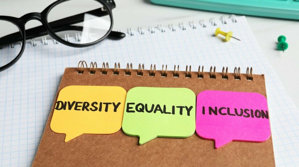  Backside of a notepad with three colorful post-its displaying the words "Diversity, Equality, and Inclusion." Next to it are a pair of black-rimmed glasses, a turquoise keyboard, and two push pins in green and yellow.