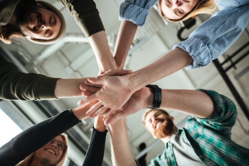 A young team is smiling and placing their hands together in a circle. The image is symbolically used to represent the work culture at weba and career entry in Steyr.