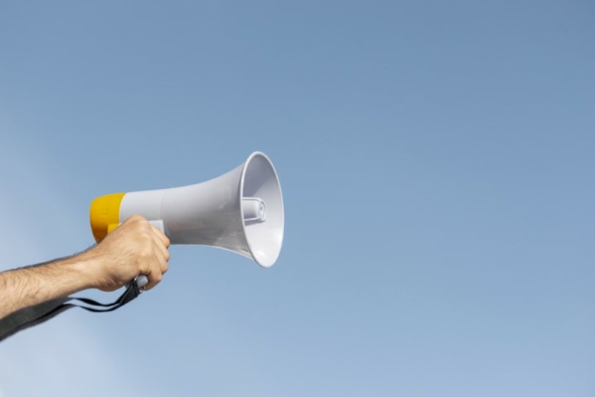 A hand holds a megaphone in the air against a blue, cloudless sky, symbolizing a guidebook for job advertisements.