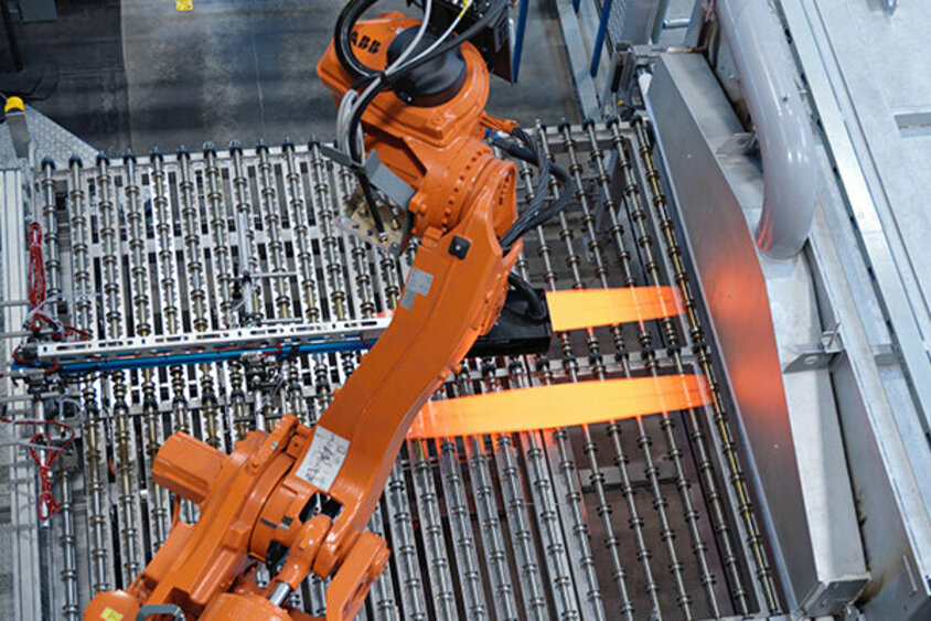 A robotic arm extracts two glowing blank plates from a continuous furnace, preparing to transfer them into a hot forming tool for shaping.