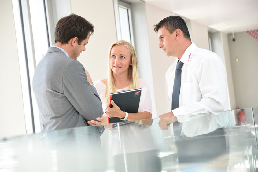 Two male employees and one female employee from weba Werkzeugbau are standing in the corridor next to a glassed area, discussing a project. A career in Steyr at weba means a shared commitment to excellence and technological leadership in forming technology.