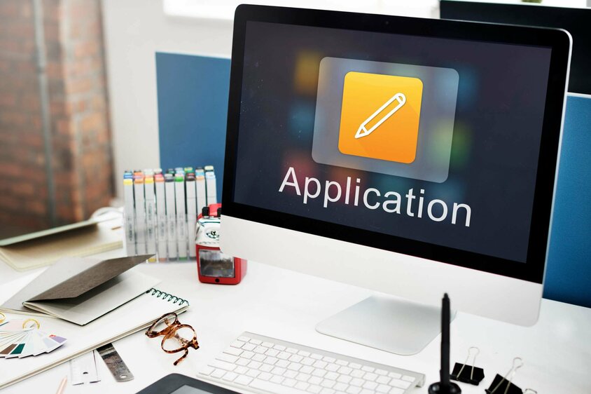 A desk with an Apple monitor, keyboard, and office supplies. The screen displays the word "Application," symbolizing job openings in Steyr at weba Werkzeugbau.