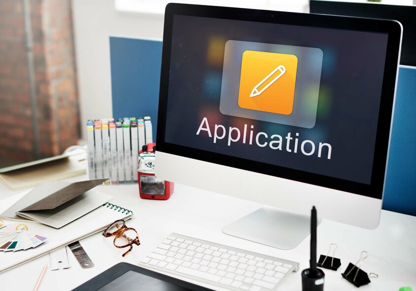 Desk with Apple screen, keyboard and office supplies. The word “Application” can be seen on the screen, symbolizing job vacancies in Steyr at weba Werkzeugbau.