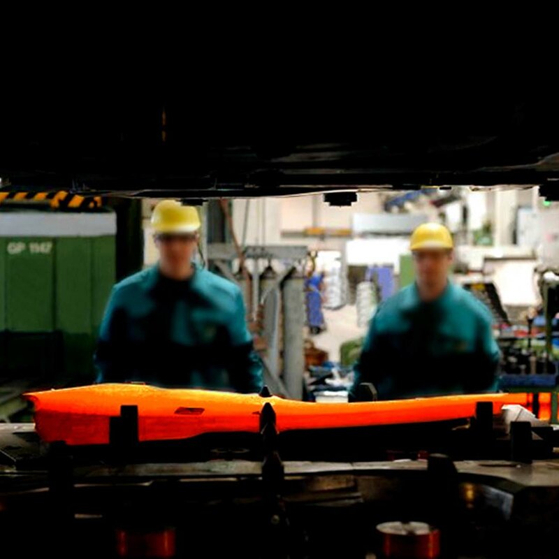 An image taken through the gap between the upper and lower part of a forming press focuses on a glowing blank inserted into a hot forming tool. Two toolmaking technicians and the blurred production can be seen in the background.