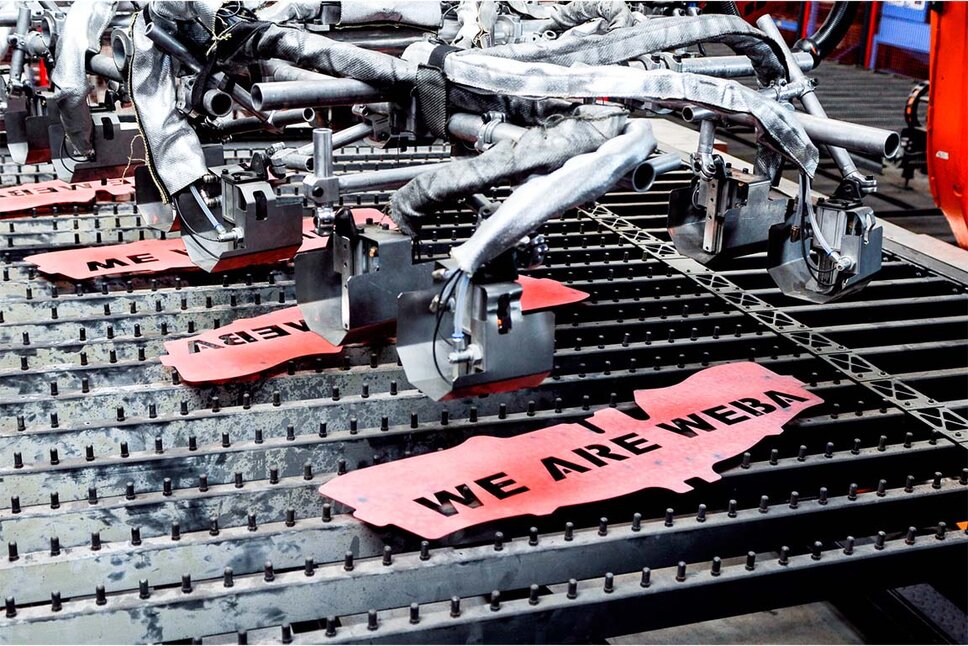 Four brightly glowing forming plates, with "WE ARE WEBA" laser-etched into them, are being carefully lifted from the table by a transfer gripper.