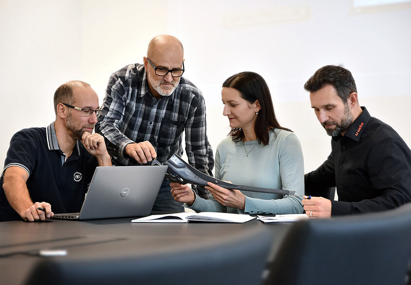 Four people are sitting at a meeting table: one person is working on a laptop, another is carefully reviewing documents, while two others are having an animated discussion about a stamped part. The scene depicts a focused and dedicated work environment. Experienced professionals can find their dream job at weba in Steyr.