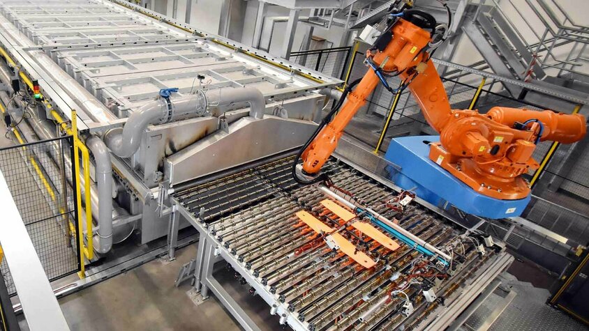 Ein orangefarbener Industrieroboter entnimmt Bauteile aus einer Warmumformungsanlage. Die Anlage ist mit komplexen Fördermechanismen und Rohrleitungen ausgestattet und befindet sich in einer modernen Produktionshalle.