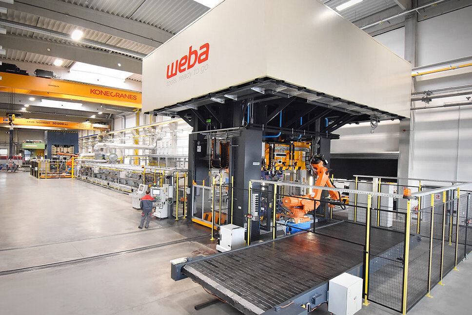 View of a series production line for hot forming, focusing on the forming press and the conveyor belt transporting finished components. A press operator is positioned next to the control panel, concentrating on monitoring the process. In the background, a portion of the roller hearth furnace is visible, used for heating the metal parts before forming. The forming press is utilized for both series production and tool tryouts.