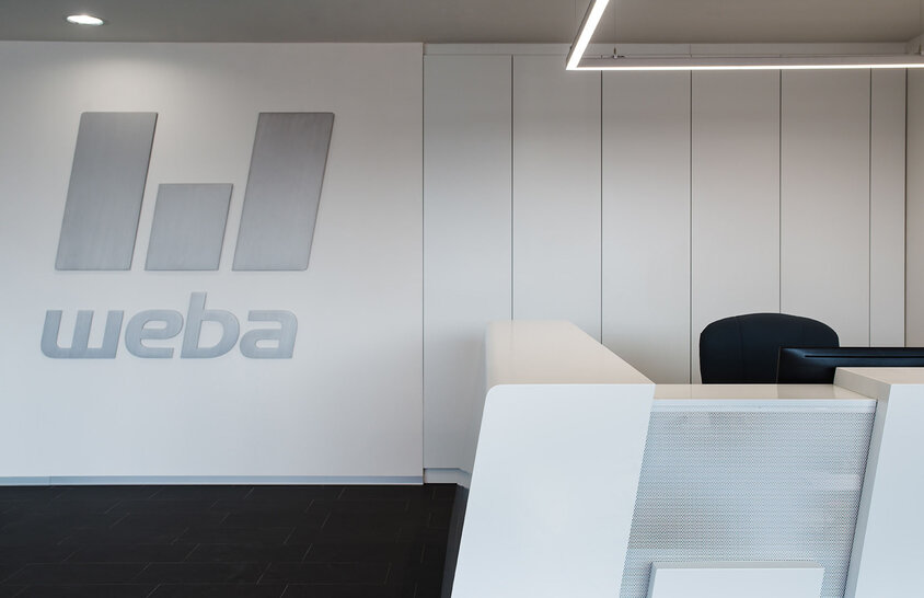 Reception area of weba Olomouc with a white reception desk and shelves. The logo in 3D lettering is in gray on the wall.