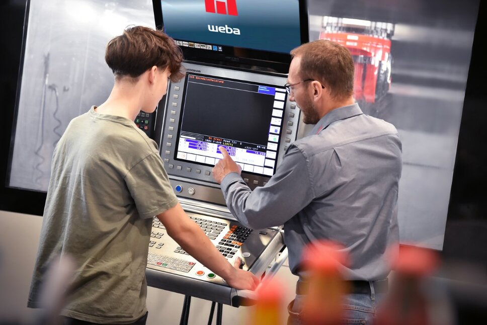 An instructor explains the operating functions of a machining center control panel to a machining technology apprentice. The apprenticeship at weba Werkzeugbau in Steyr focuses on hands-on training directly on the machines.