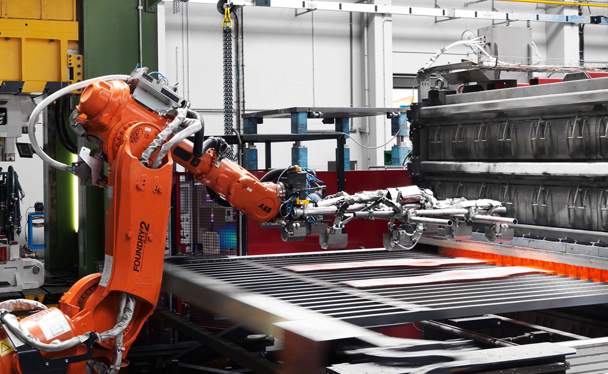  Ein Robotertransferarm greift zwei rotglühende Metallplatinen aus dem Inneren eines Kammerofens, um sie in ein Warmumformwerkzeug für die Herstellung von Strukturbauteilen zu transferieren. Die leuchtend roten Platinen stehen im Kontrast zum metallischen Glanz des Roboterarms und der dunkleren Umgebung des Industrieofens. Diese Szene veranschaulicht die Präzision und Automatisierung in modernen Fertigungsprozessen, insbesondere in der Herstellung hochfester Metallkomponenten mittels Warmumformung für den Einsatz in anspruchsvollen Anwendungen.